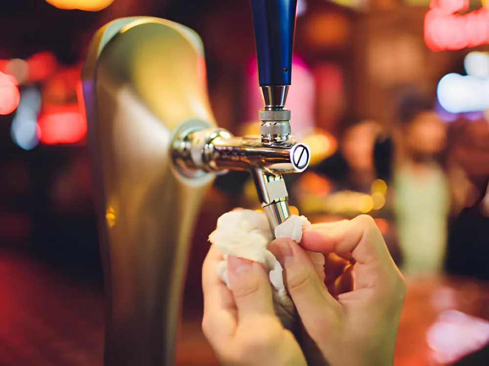 A cloth wiping a single draft faucet, bar lights behind